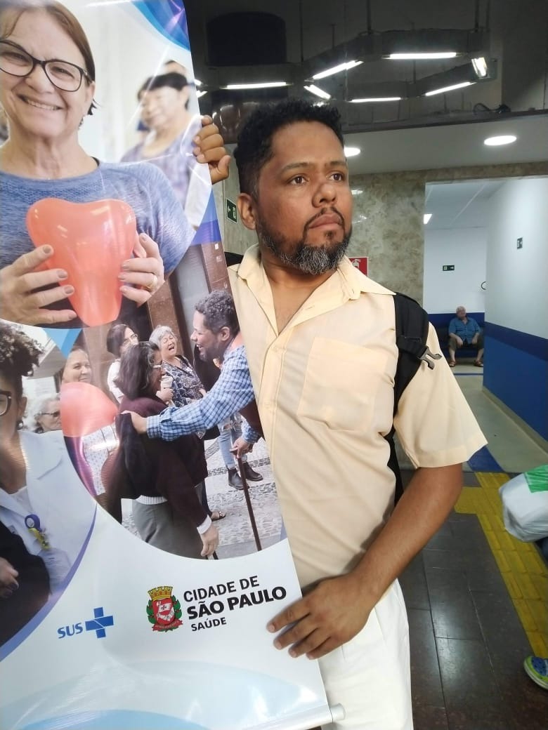 A foto mostra Edson segurando um banner que contém várias imagens de profissionais de saúde interagindo com o público; na parte inferior do banner está a logomarca da Secretaria de Saúde do Município de São Paulo